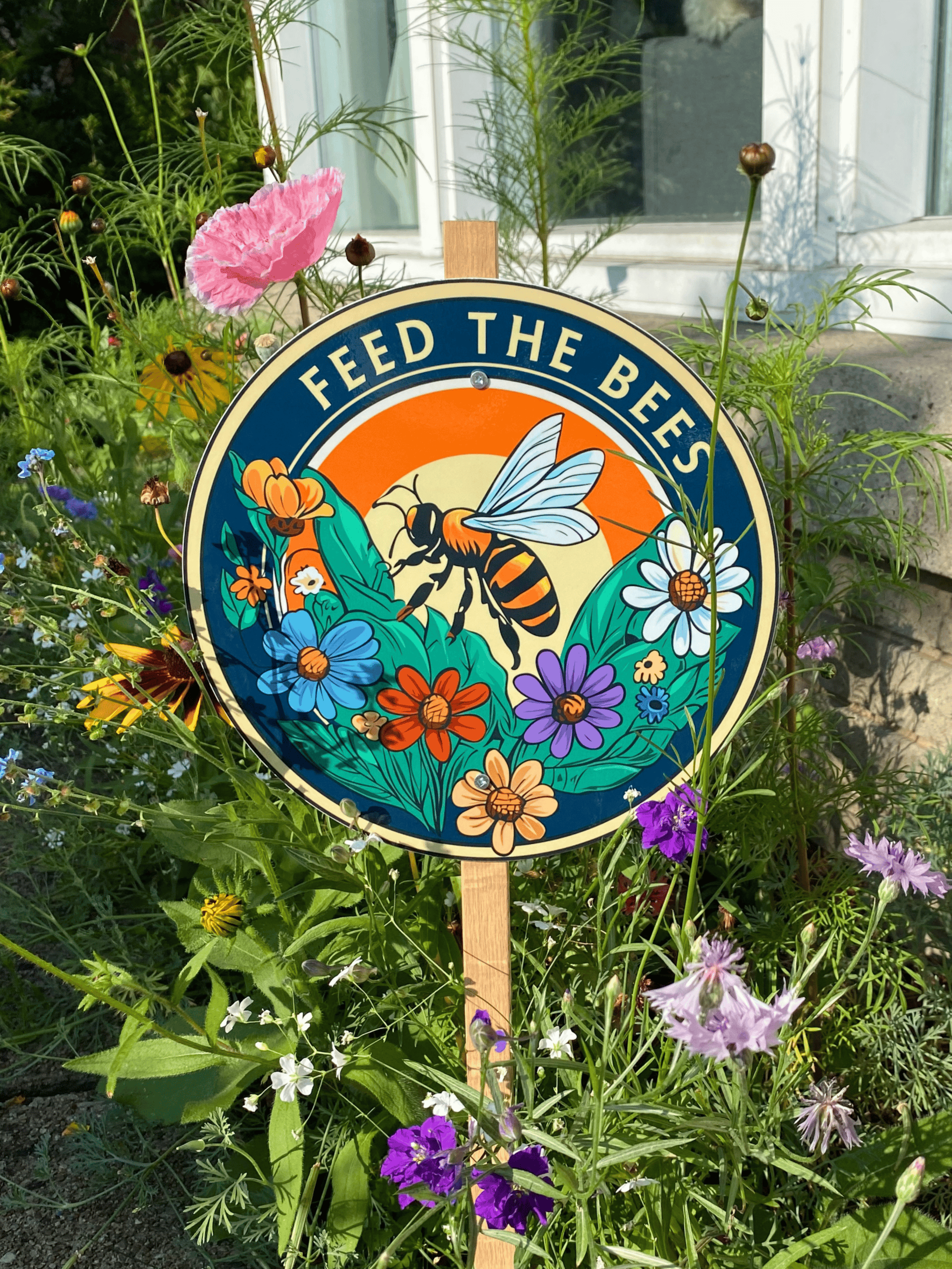 Feed the Bees hand-illustrated garden sign displayed among red zinnias
